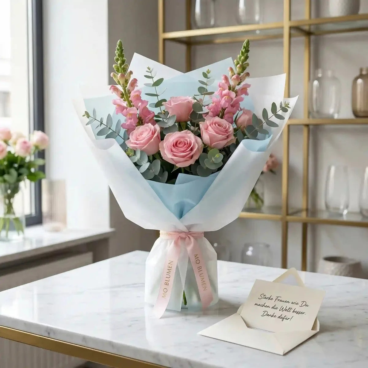Ein Blumenstrauß mit zarten rosa Rosen von MO BLUMEN, mit rosa Löwenmäulchen, Eukalyptus und in hellblau-weißem Papier gebundenem rosa Band. Daneben liegt ein Notizzettel.